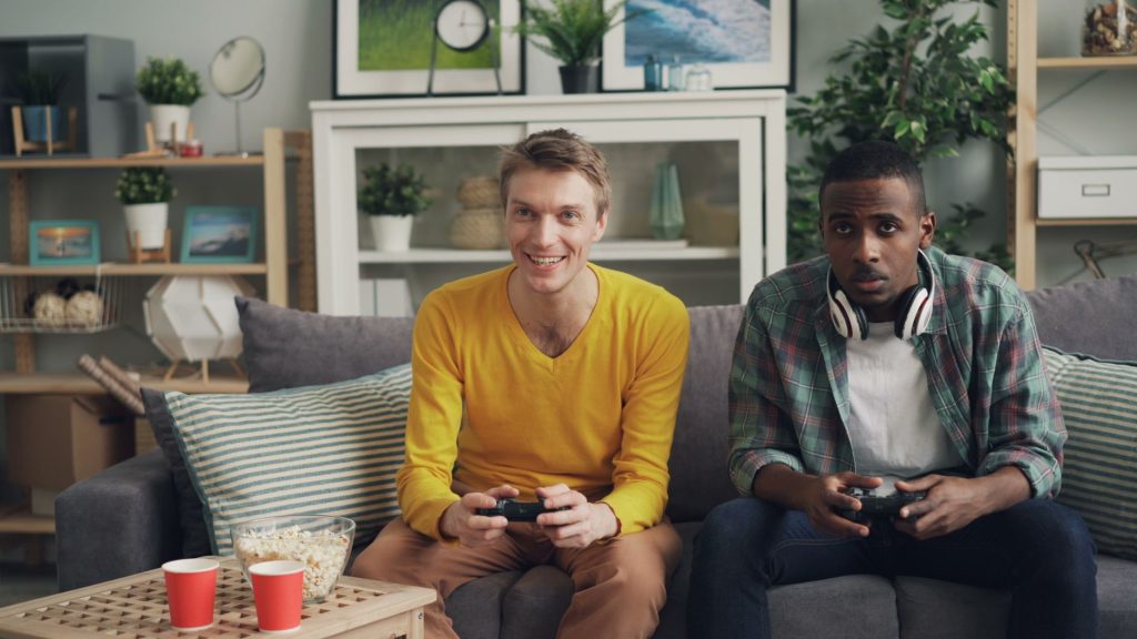 Two friends playing video games together on a sofa, one holding a controller and the other wearing headphones. — Official UFA