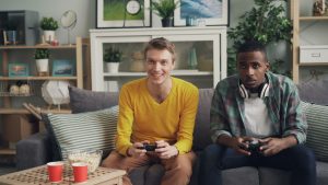 Two friends playing video games together on a sofa, one holding a controller and the other wearing headphones. — Official UFA